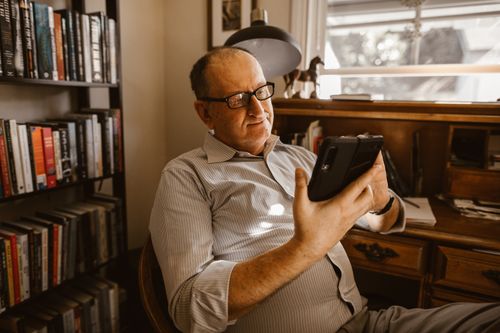 Free Elderly man using a tablet indoors surrounded by books, showcasing technology engagement. Stock Photo