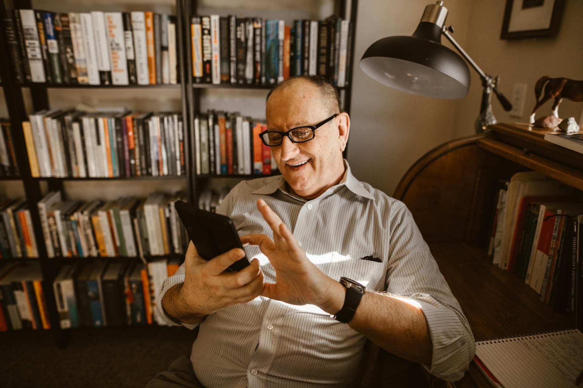 Adult man with eyeglasses smiling at his smartphone in a library with bookshelves and warm lighting.