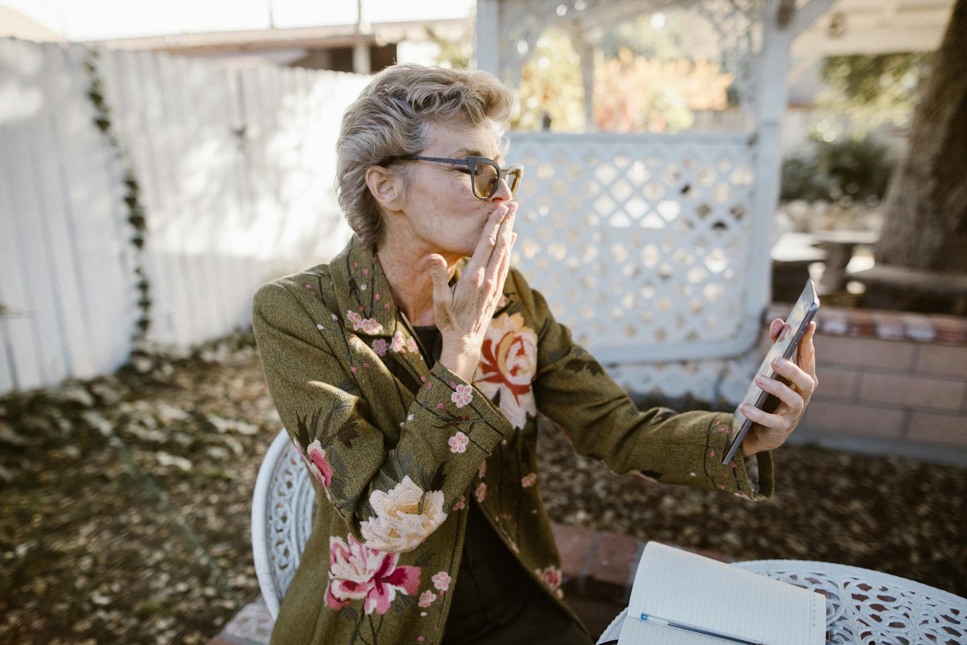 A cheerful senior woman using a smartphone for a video call, blowing a kiss in an outdoor garden setting.