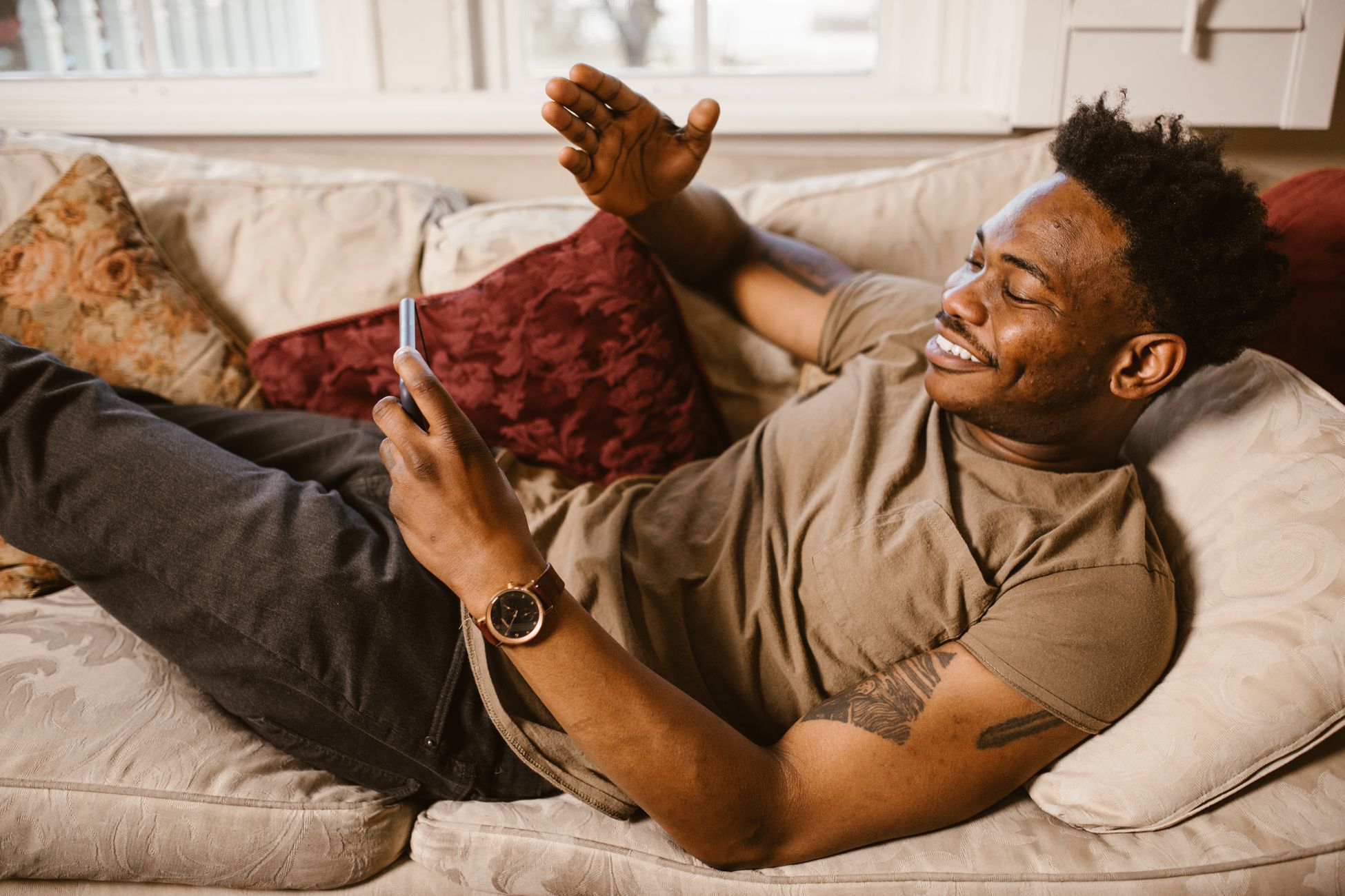 African American man lying on couch, smiling and using smartphone for communication.