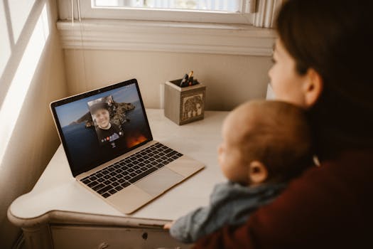 A mother holding her baby while making a video call on a laptop indoors.