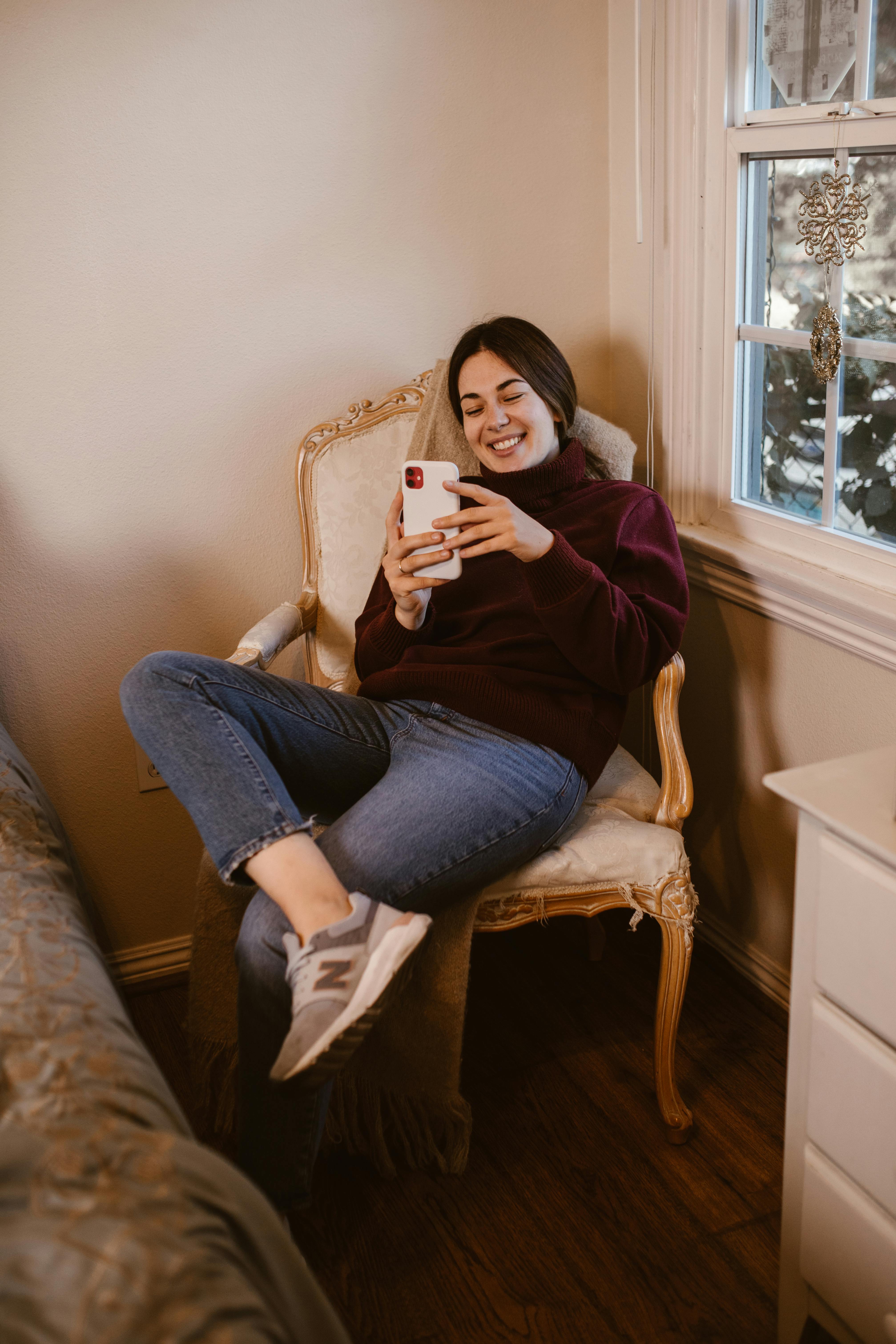 Free A young woman smiling while using a smartphone in a cozy indoor setting. Stock Photo
