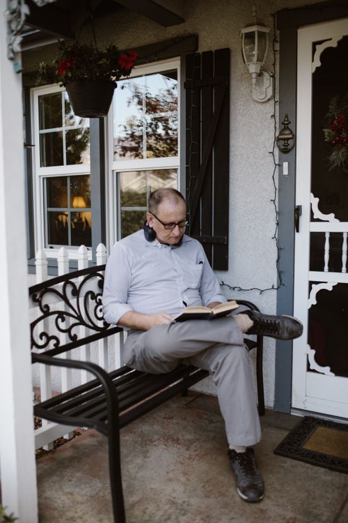 Free Elderly man sitting on porch bench reading a book outdoors, encapsulating leisure and relaxation. Stock Photo