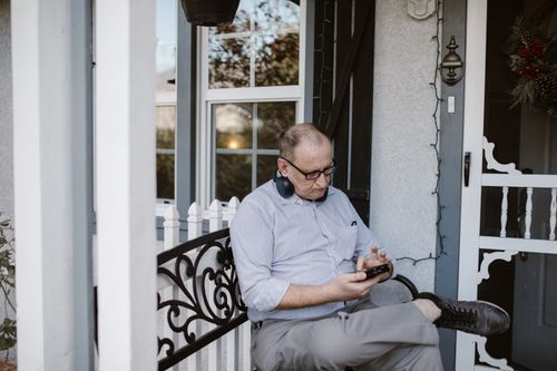 Free Elderly man with headphones browsing phone while sitting on a porch bench. Stock Photo