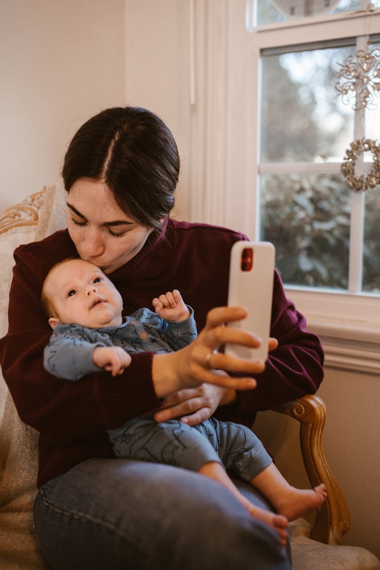 A Mother Kissing Her Baby While Holding Her Mobile Phone