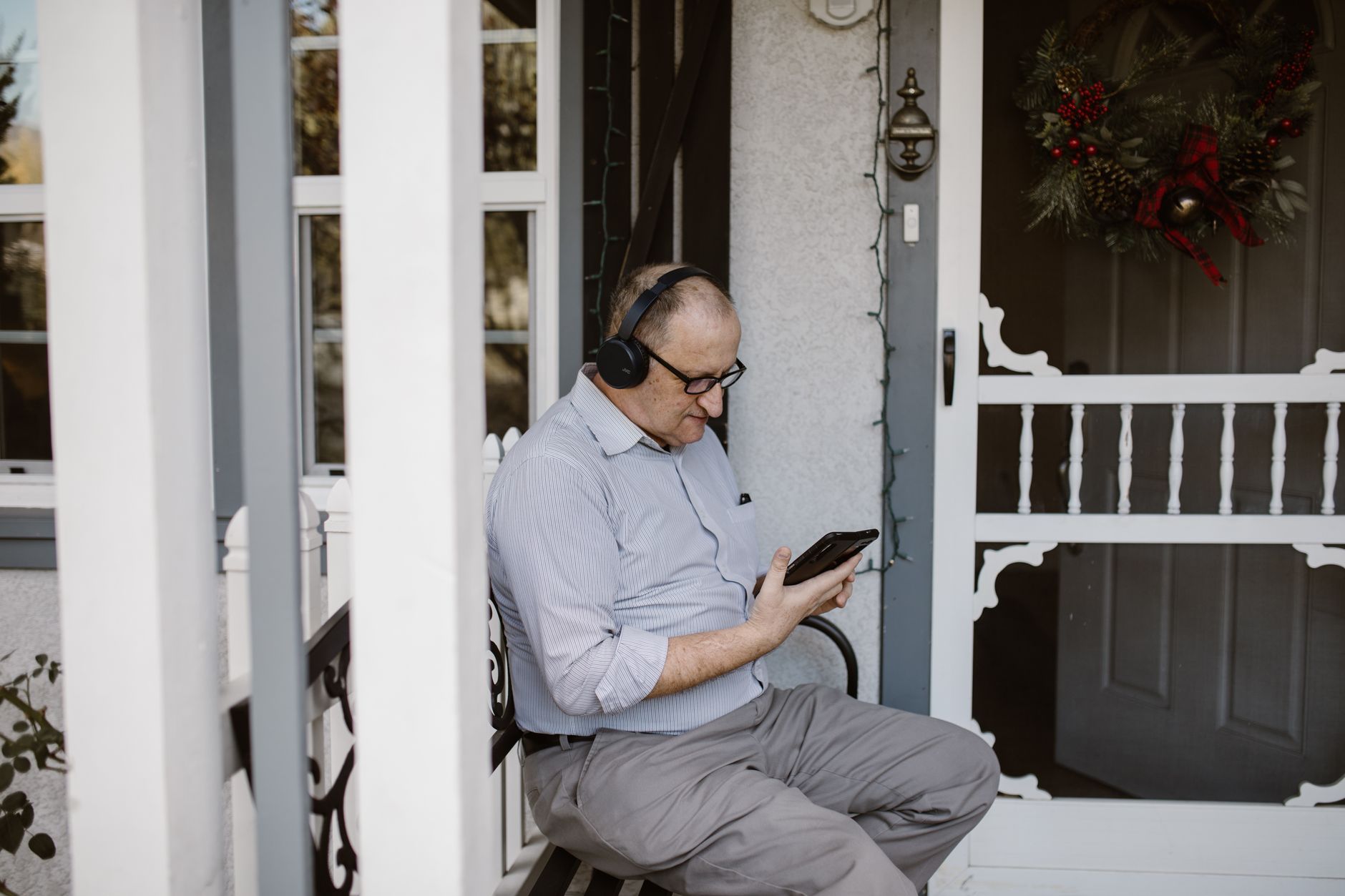 경기도 어르신 스마트폰 무료 교육 2 Elderly man using smartphone and headphones on porch. Winter decor suggests festive mood.