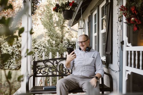 Free Senior man using phone and headphones for video call on home porch. Stock Photo