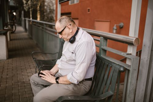 Free Senior man sitting on an outdoor bench with headphones, using a smartphone. Stock Photo