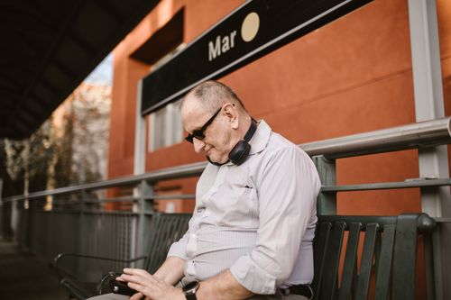 Free Elderly man sitting on a bench at a train station listening to music with headphones. Stock Photo