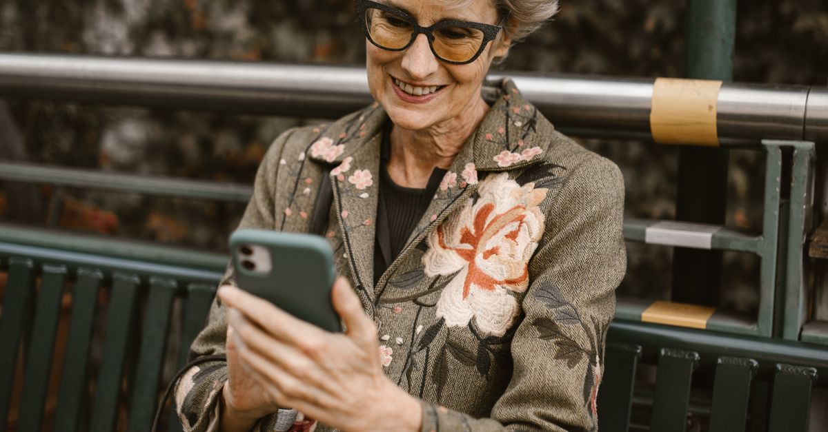 Rüyada Uzaktan Aşk Yaşamak - Rüya Tabiri ve Anlamı - A fashionable senior woman wearing glasses enjoys her phone while sitting on a park bench during autumn.