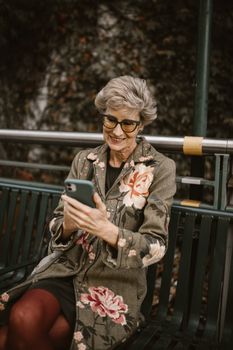 A fashionable senior woman wearing glasses enjoys her phone while sitting on a park bench during autumn.