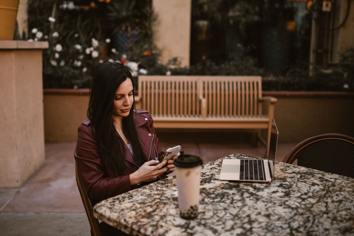 Femeie la o masă de cafenea în aer liber, cu telefonul, cafeaua și laptopul în față