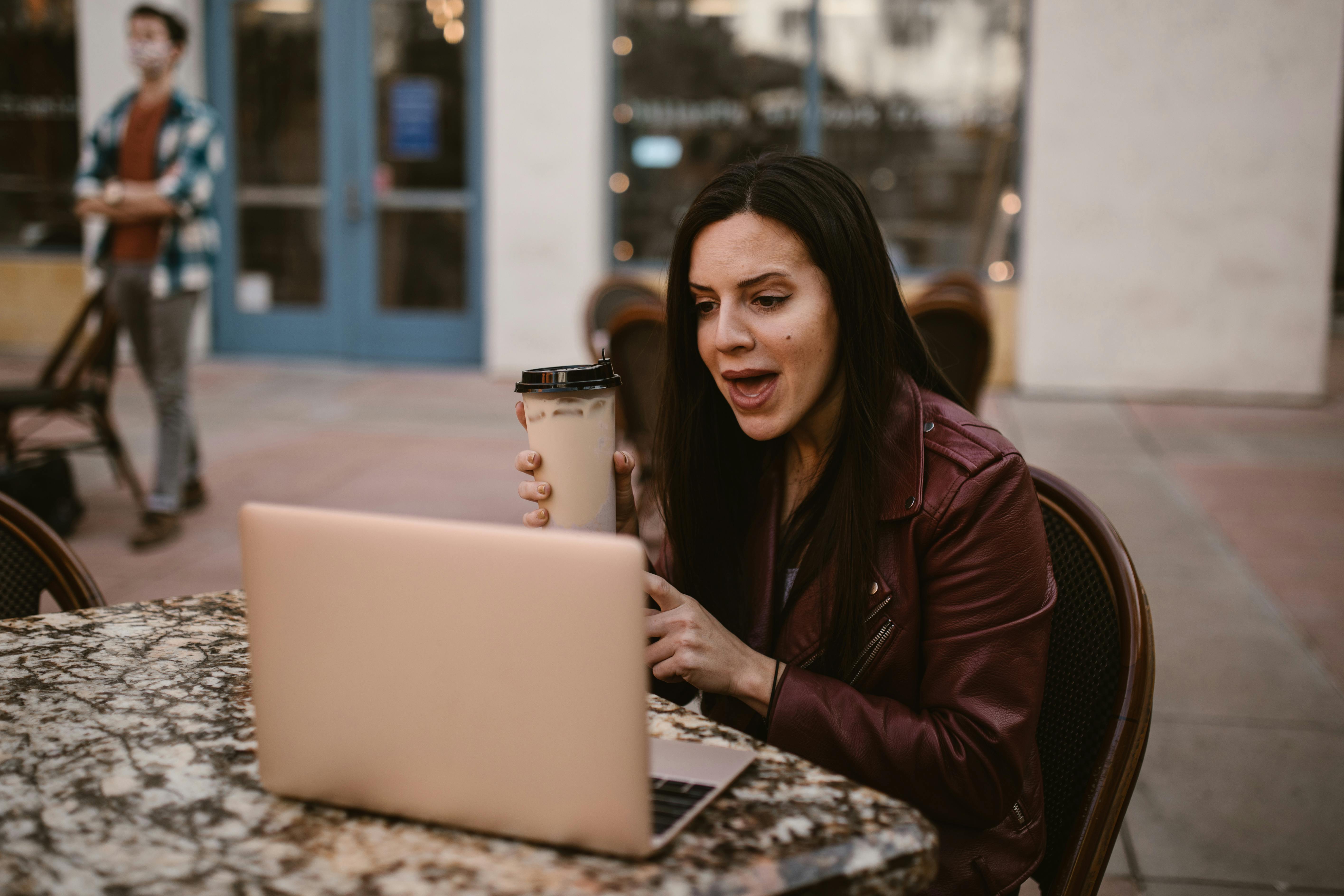 a woman in brown leather jacket holding a disposable cup while talking in front of her laptop