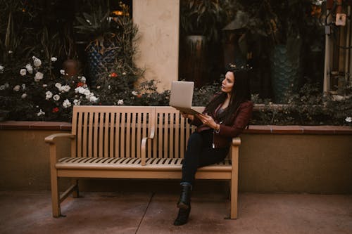 Женщина в коричневой куртке и черных штанах сидит на деревянной скамейке с ноутбуком