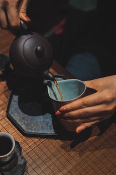 Close-up of a tea pot pouring tea into a porcelain cup on a wooden mat.