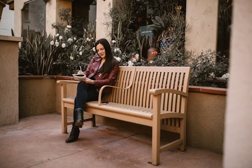 Женщина в коричневой куртке сидит на коричневой деревянной скамейке и пишет в блокноте
