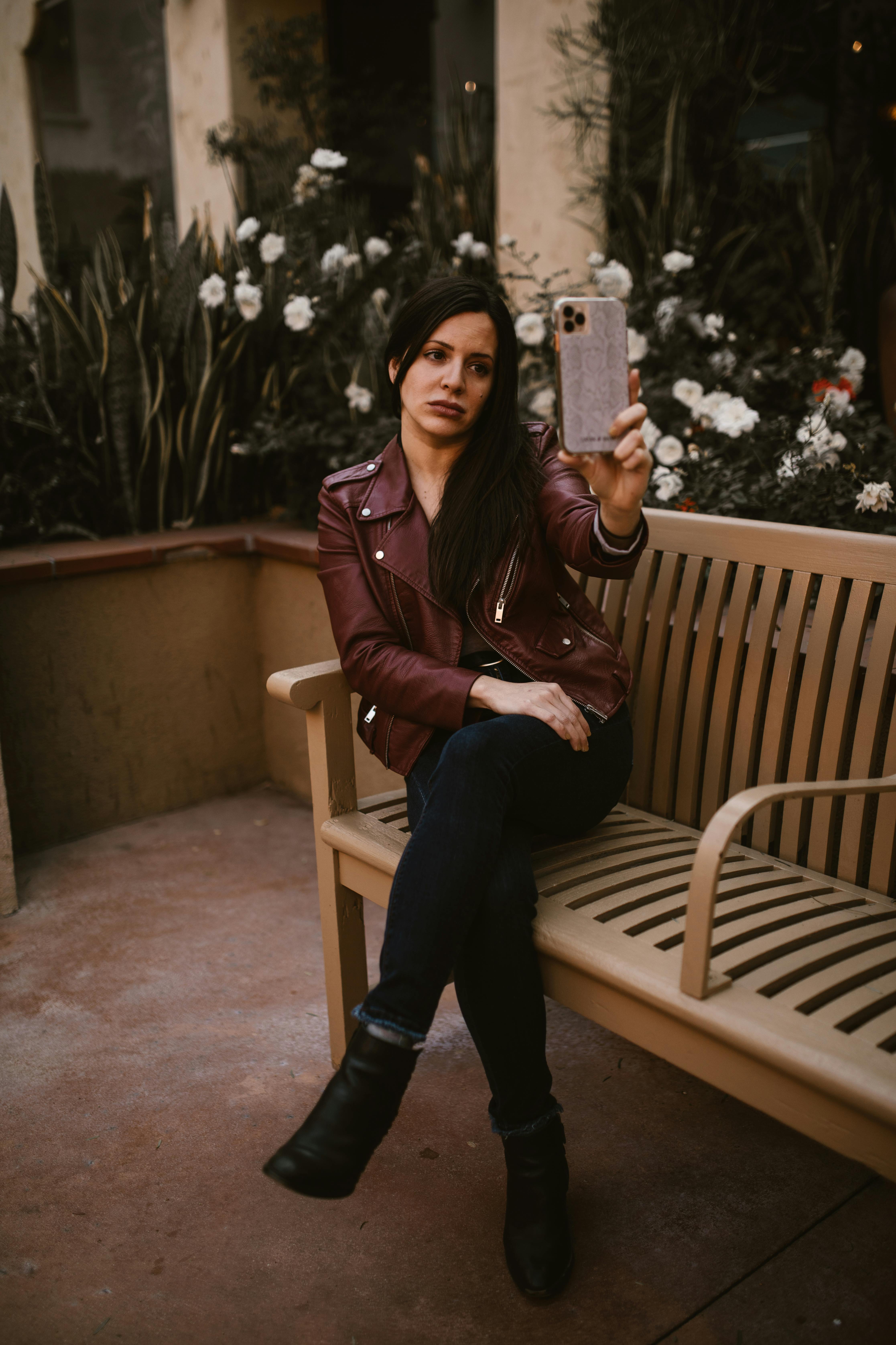 a woman in brown leather jacket sitting on the bench while holding her mobile phone