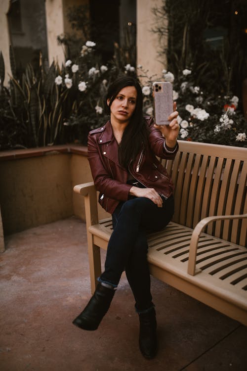 A Woman in Brown Leather Jacket Sitting on the Bench while Holding Her Mobile Phone