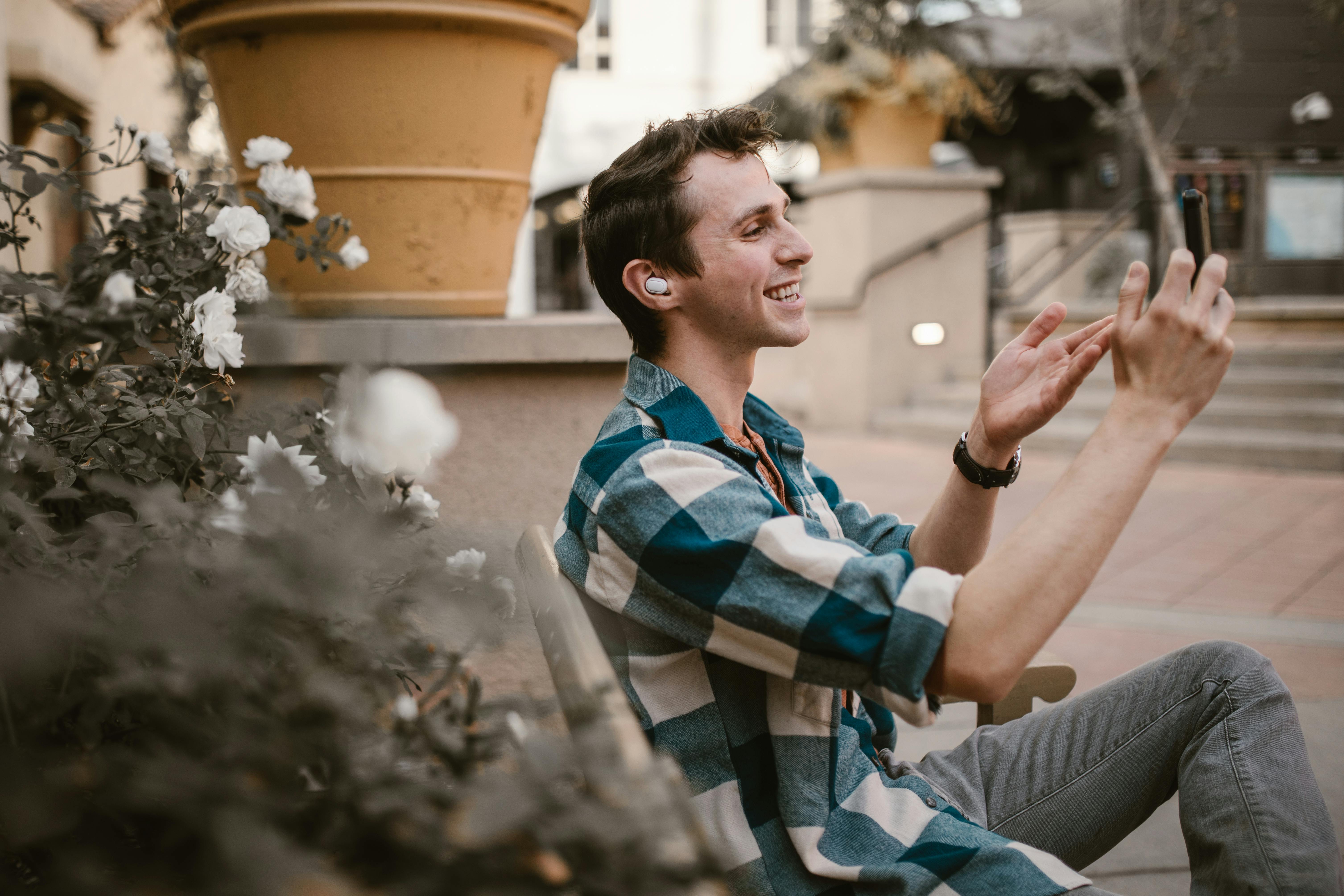 wireless earbuds with long battery life uk - Smiling man using smartphone for video call while sitting outdoors in park.