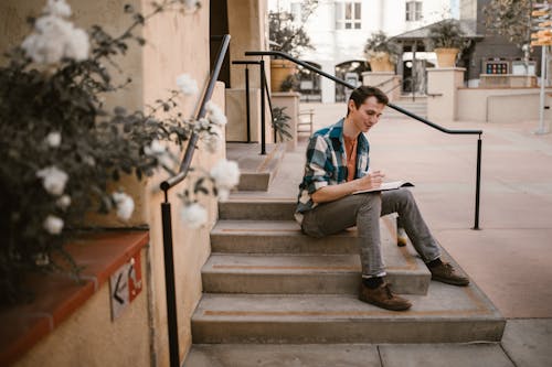 A Man Sitting on a Concrete Stairs while Writing on His Notebook