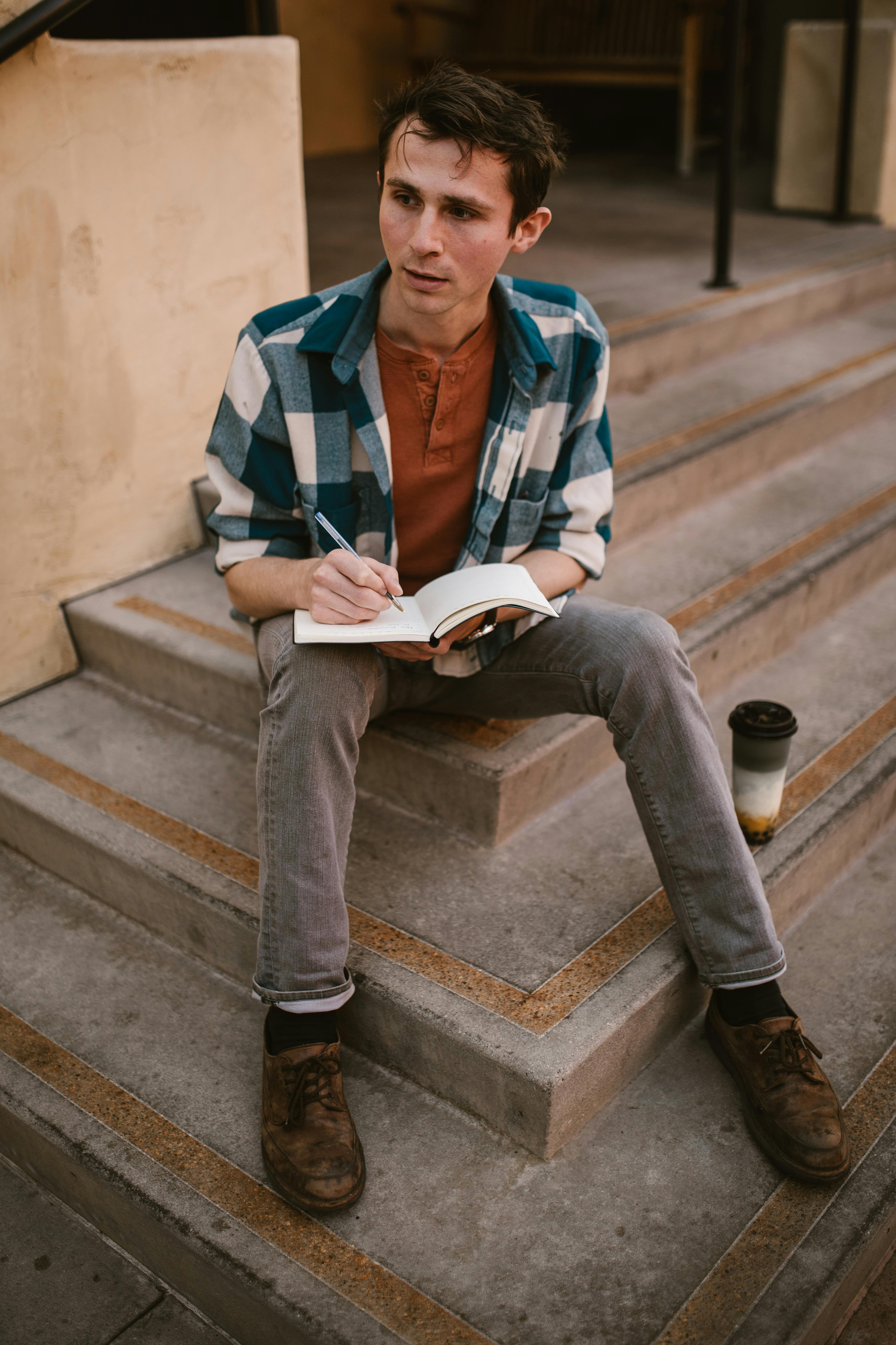 Casually dressed man writing in a notebook while seated on steps outdoors with a coffee cup nearby.