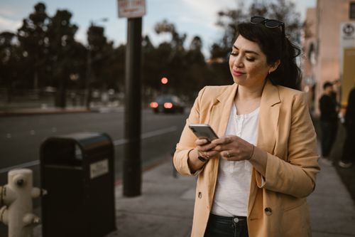 Free Casual woman in beige blazer on city street using smartphone, smiling at sunset. Stock Photo