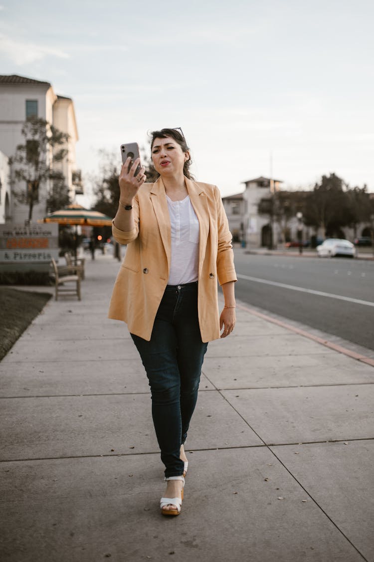 A Woman In Beige Blazer And Black Pants Walking On The Street While Talking On The Phone