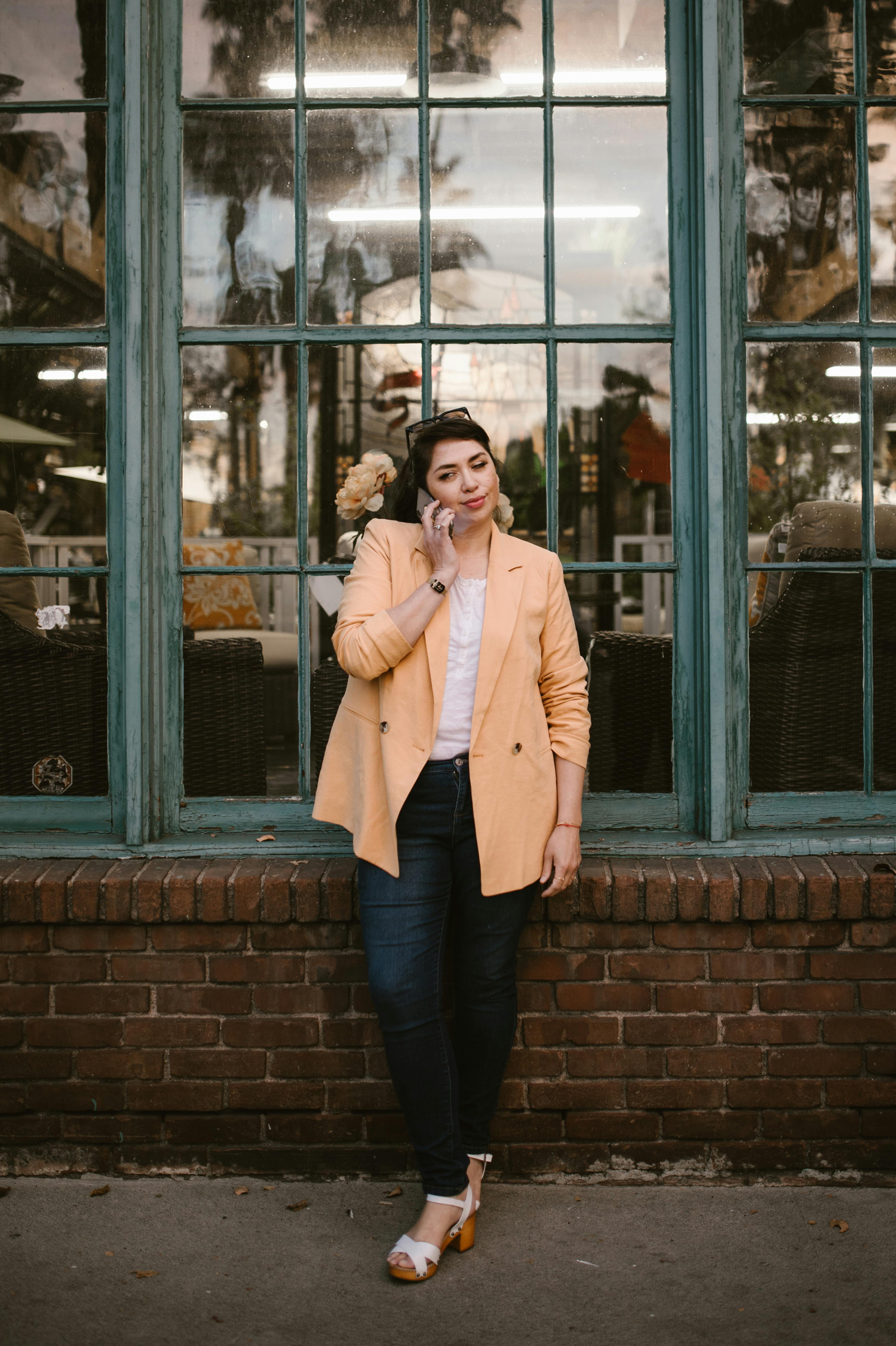 a woman in beige coat standing near the window while talking on the phone