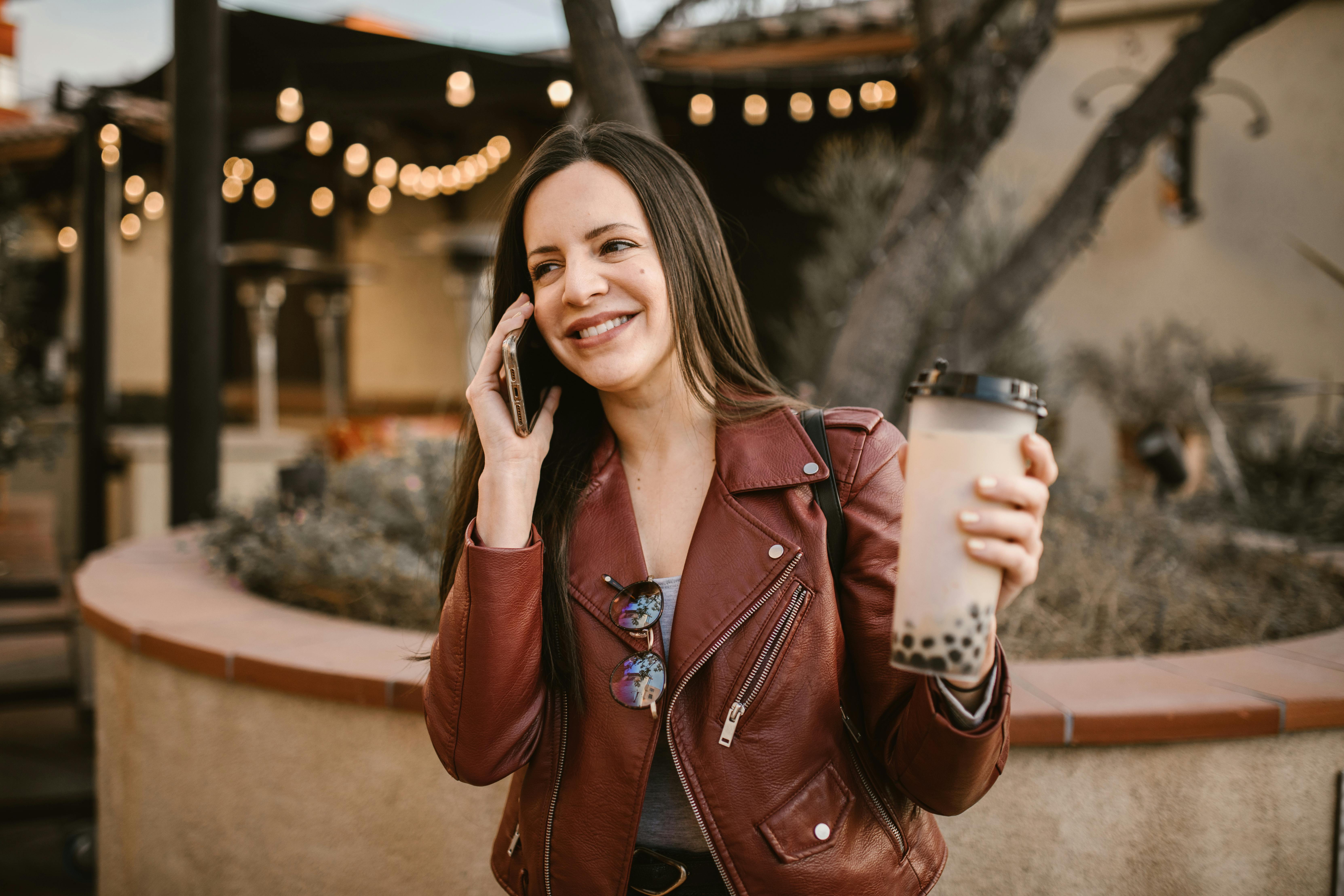 a woman in brown leather jacket talking on the phone while holding her drink