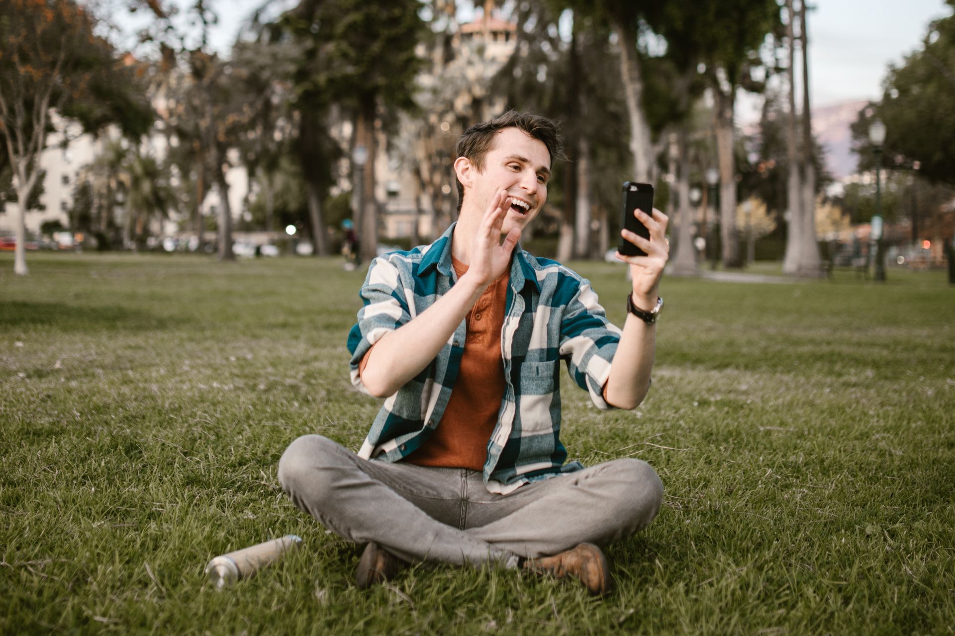 A cheerful man video calls using his smartphone while sitting on a park lawn.