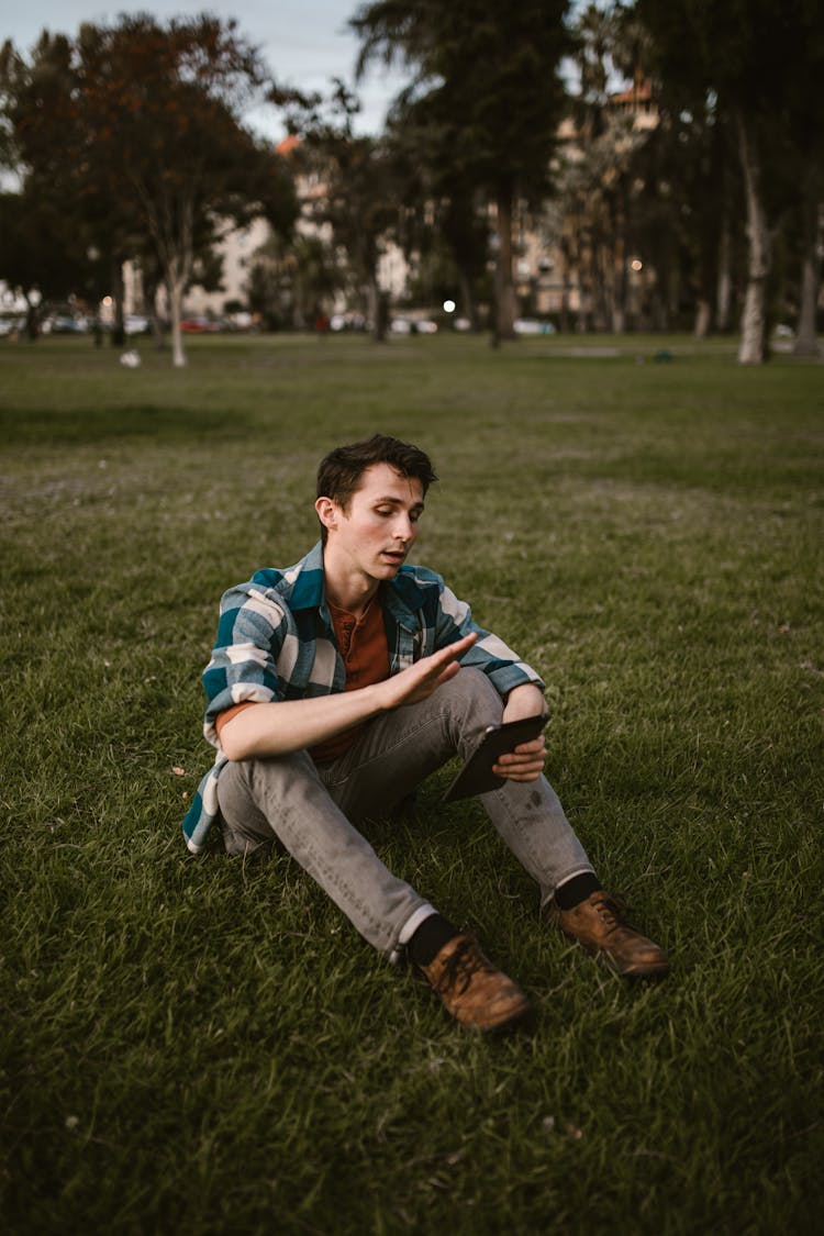 A Man Sitting On Grass Field While Using His Tablet