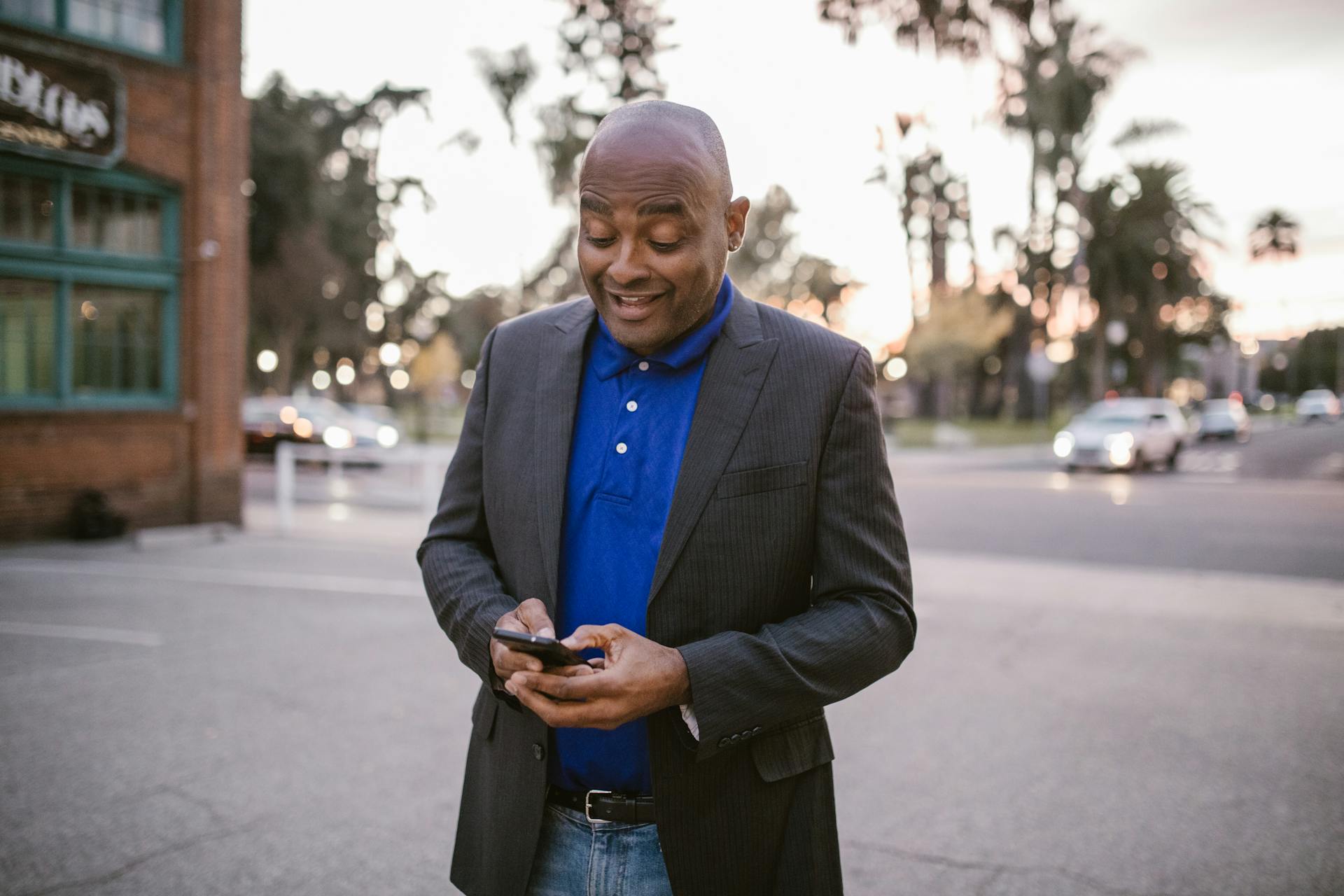 An adult man in a suit is happily using his smartphone outside during sunset, showcasing communication and modern lifestyle.