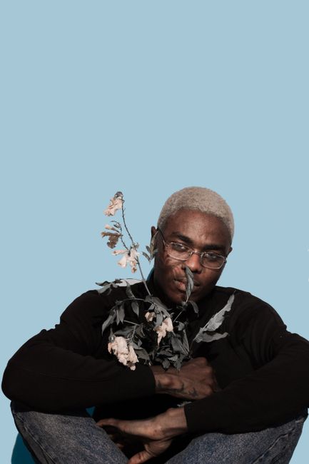Calm young black male millennial with dyed hair in stylish clothes and eyeglasses sitting near blue wall with bunch of withered flowers and looking at camera