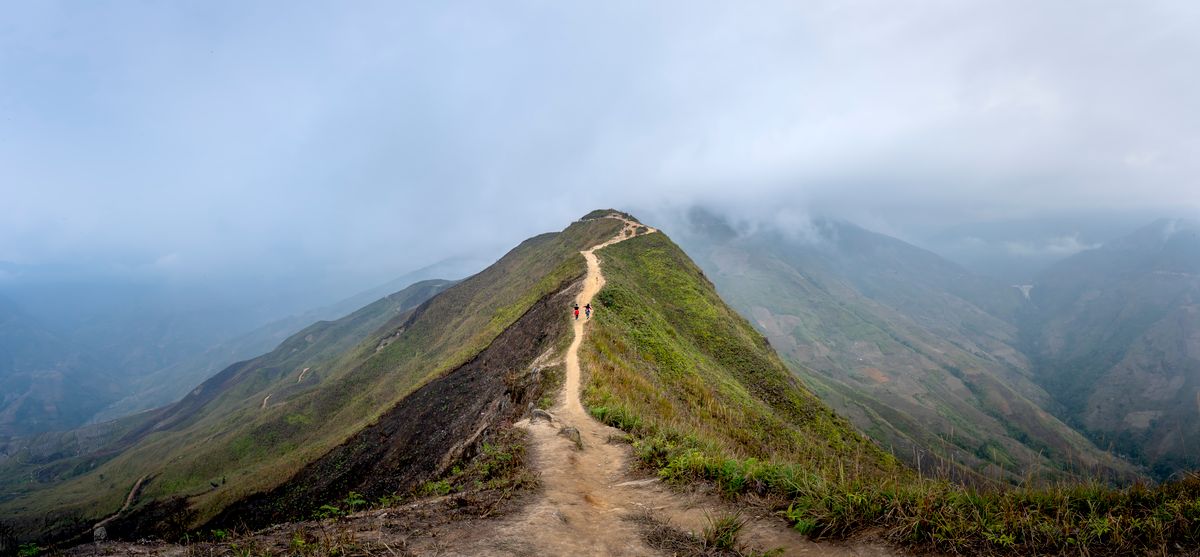 Hikers strung along a narrow grass ridge trail, misty forested valleys falling away on both sides of the crest.