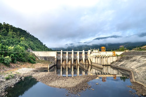 Industrial hydroelectric power plant located near river and stony coast against green mountain range covered with mist on summer day