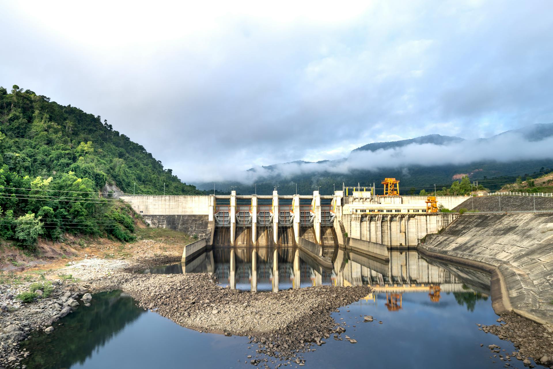 Industrial hydroelectric power plant located near river and stony coast against green mountain range covered with mist on summer day