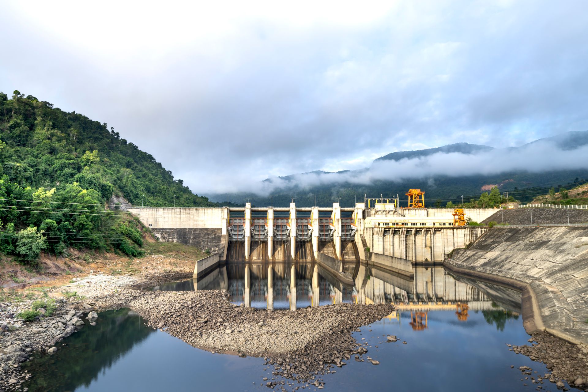Industrial hydroelectric power plant located near river and stony coast against green mountain range