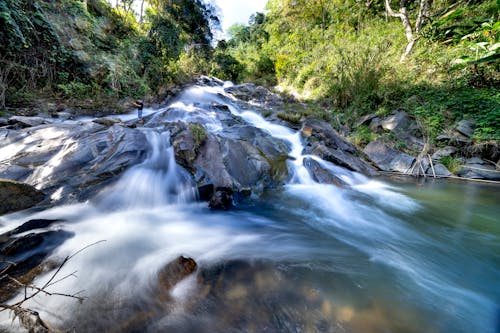 Long exposure of rapid stream flowing from rough rocky boulders into river flowing near green trees in nature on summer day