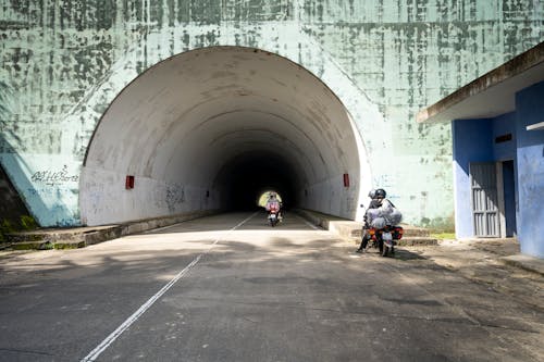 People riding motorbikes on straight asphalt roadway leading to shabby tunnel near old building in suburb area of sunny countryside