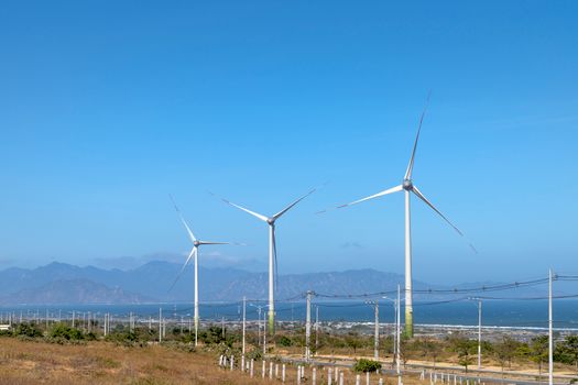 Scenery view of wind turbines in row on terrain with plants against ocean and mounts in daytime
