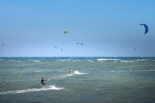Anonymous sportspeople with power kites practicing kitesurfing on wavy ocean under blue sky on stormy weather