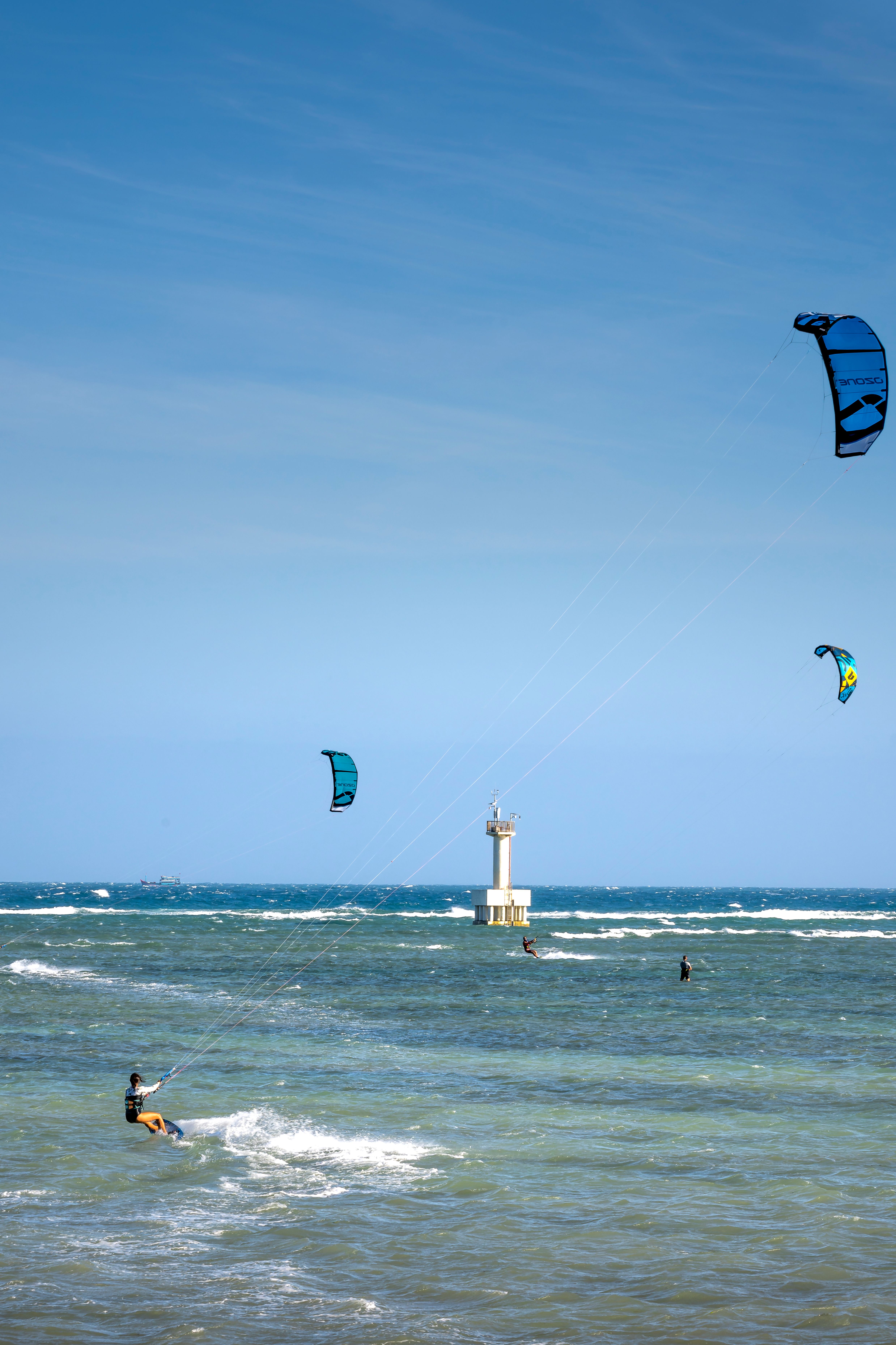 Unrecognizable sportspeople with power kites practicing kiteboarding on foamy ocean with lighthouse under cloudy blue sky