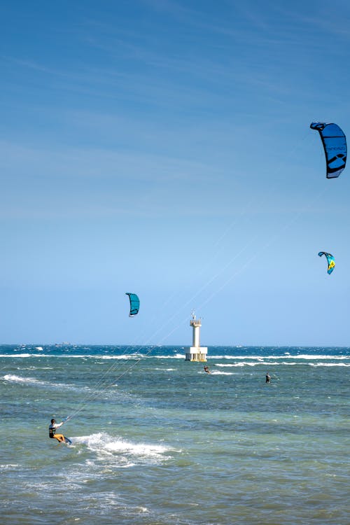 Free Unrecognizable sportspeople with power kites practicing kiteboarding on foamy ocean with lighthouse under cloudy blue sky Stock Photo