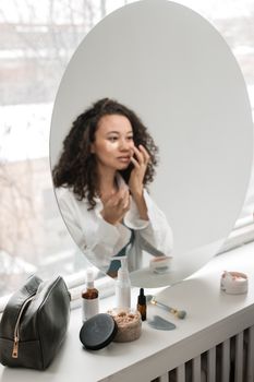 A woman with curly hair applies skincare products in front of a round mirror.