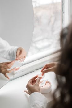A woman applies eye patches beside a window, reflecting a serene self-care moment.