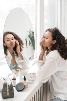 A woman applies skincare products, sitting at a vanity with a mirror in a bright room.