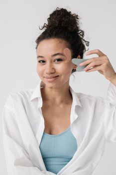 Smiling woman using a gua sha tool and eye patch for a beauty routine indoors.