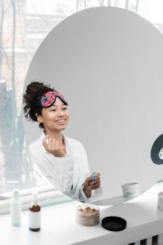 Smiling woman applies skincare products in front of a large round mirror during the day.