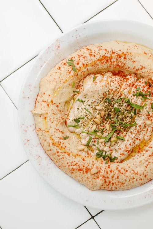 Free A close-up of authentic hummus topped with herbs, spices, and pine nuts on a white tiled surface. Stock Photo