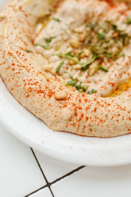 A bowl of pale, creamy hummus swirled with a spoon, pooled with olive oil in the center and dusted with smoked paprika, served with cucumber slices and pita triangles.